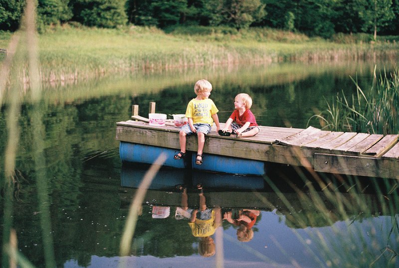 Children sitting on a wooden dock by a calm lake, enjoying a sunny day outdoors, captured with natural lighting for a timeless outdoor photography style.