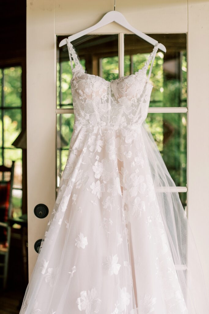 Elegant lace wedding dress hanging on a white hanger in front of a glass door with lush greenery outside, showcasing Bridal Photography, Wedding Dress, and Bridal Gown.