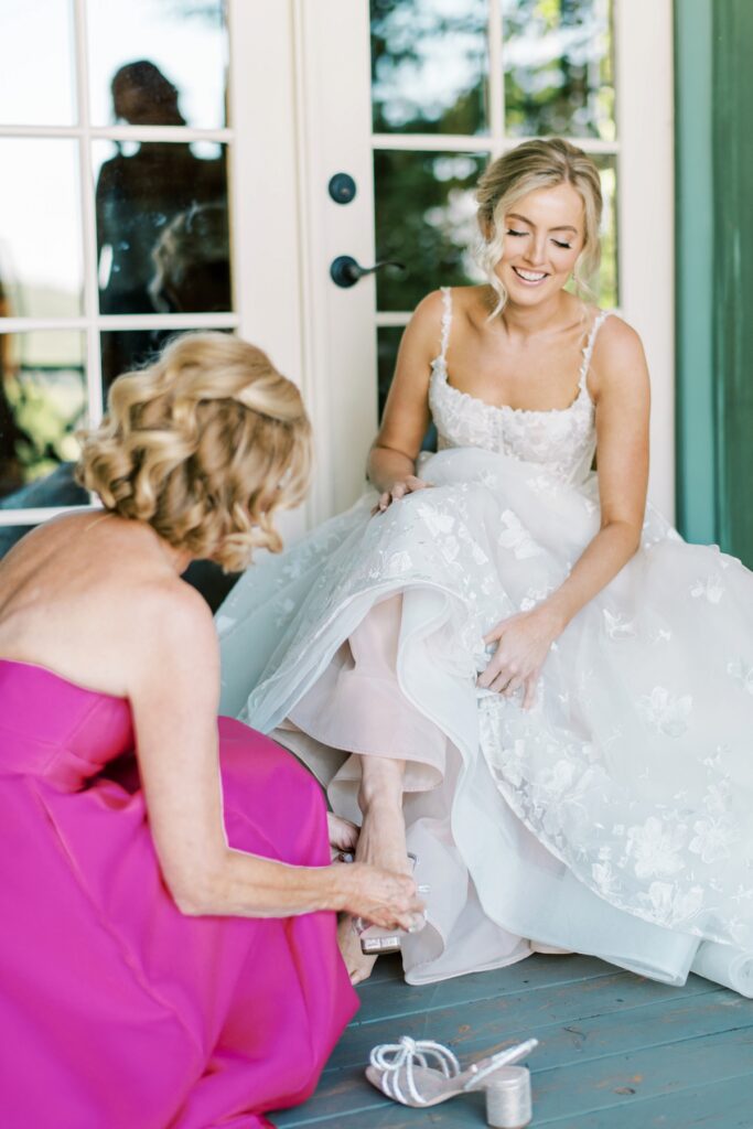 Elegant bride getting ready for wedding, being assisted with shoes by a bridesmaid, in a bright and cheerful setting with natural light, floral lace dress, and joyful atmosphere at Meghan Rose Photography.