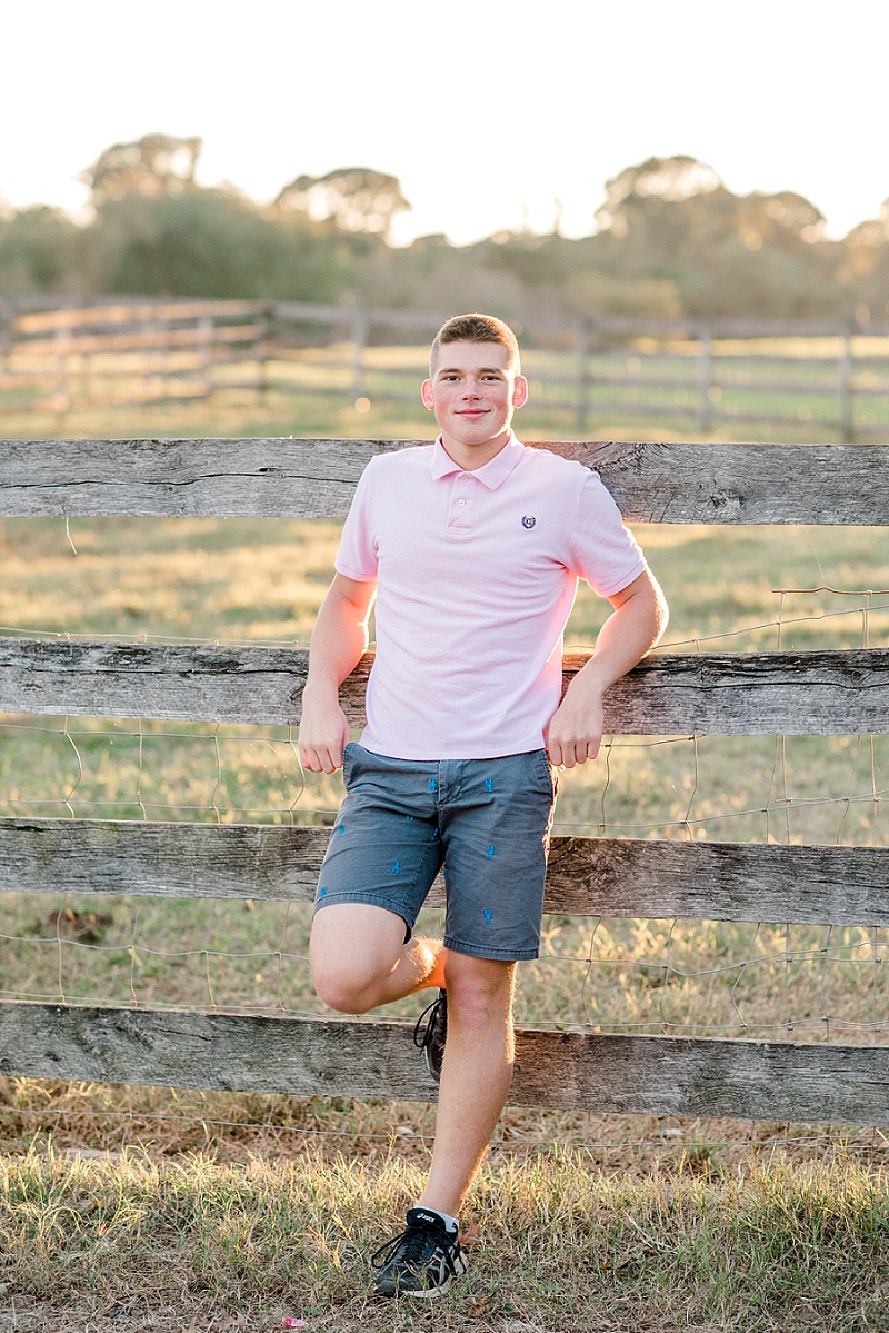 senior boy in pink shirt leaning against wooden fence