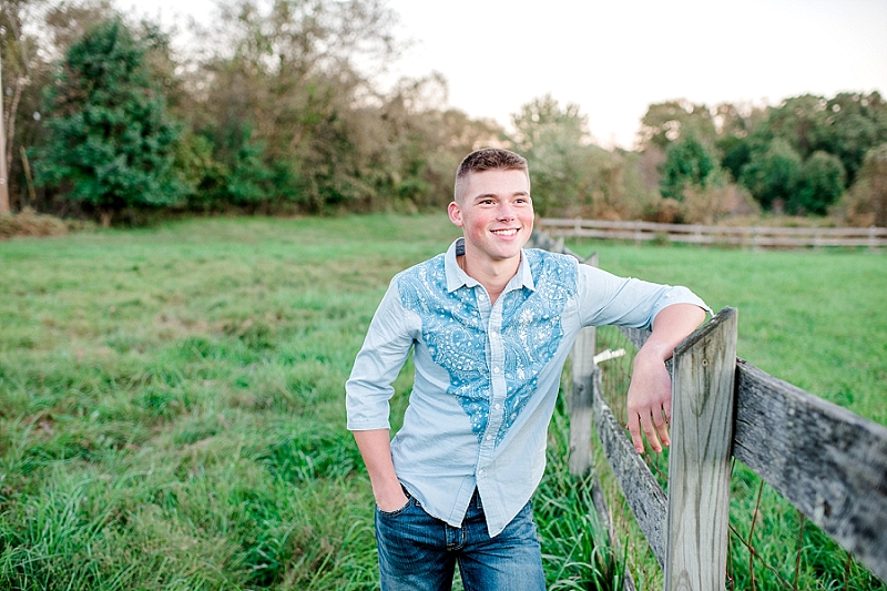 senior boy in a green field leaning against a wooden fence